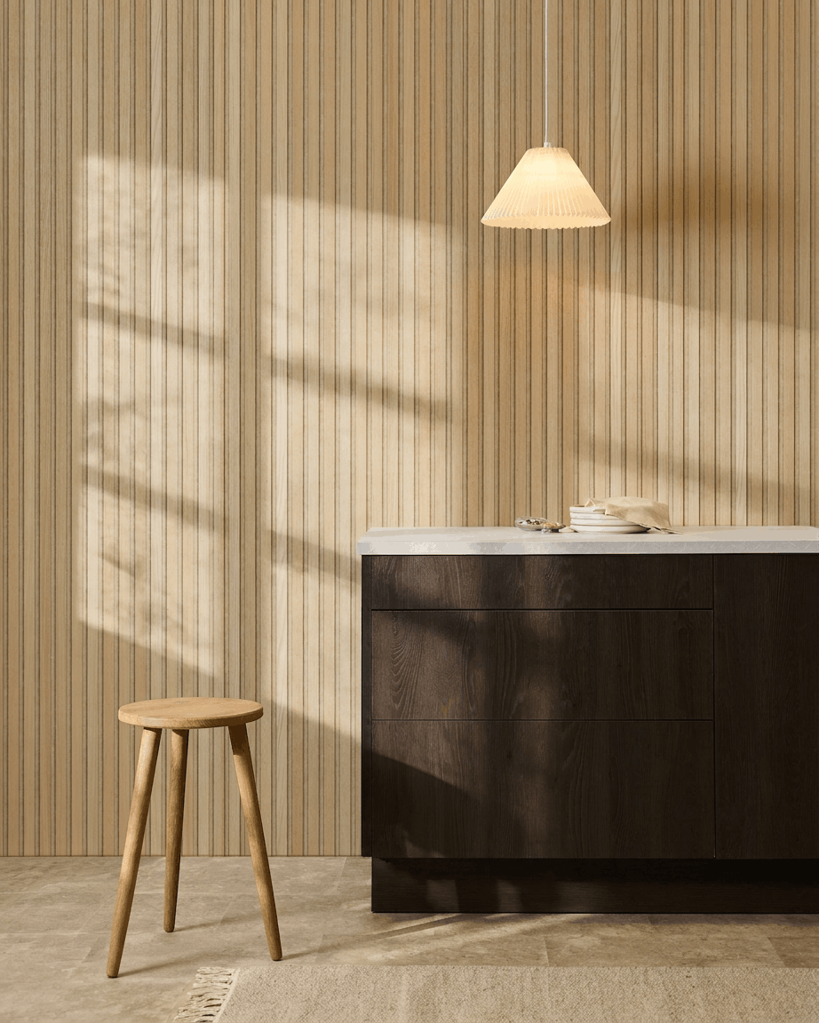 Skandinavische Küche mit WoodUpp Akupanel Classic Oak Akustik-Lamellenwandpaneel, dunklen Holzschränken, Holzstuhl, Pendelleuchte und Sonnenlicht, das Schatten wirft.