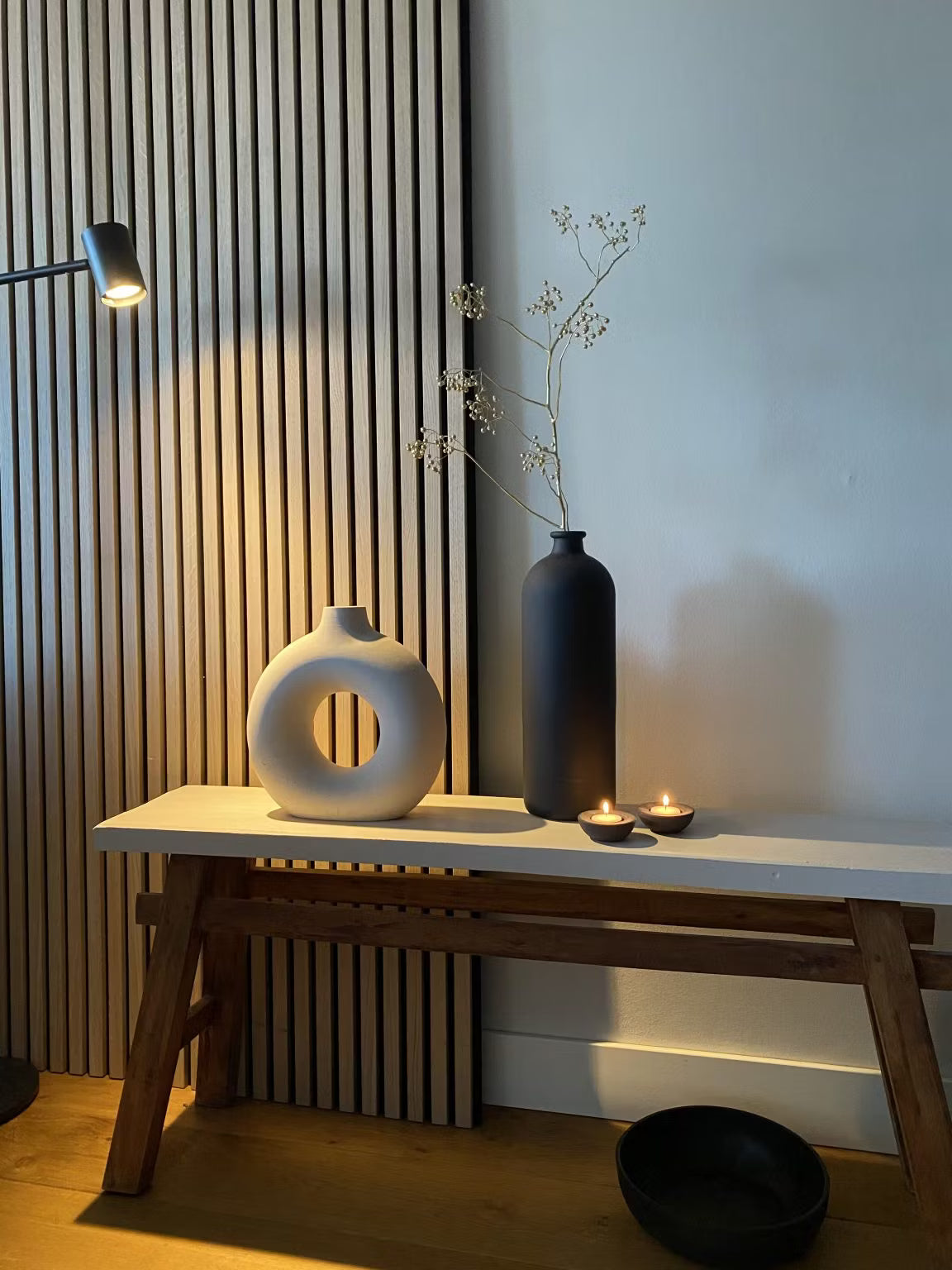 Minimalistische Dekoration mit einer weißen runden Vase, einer schwarzen hohen Vase mit getrockneten Stängeln, zwei Teelichtkerzen und einer schwarzen Schale vor dem Hintergrund einer Akupanel-Holzlatten-Akustikplatte.