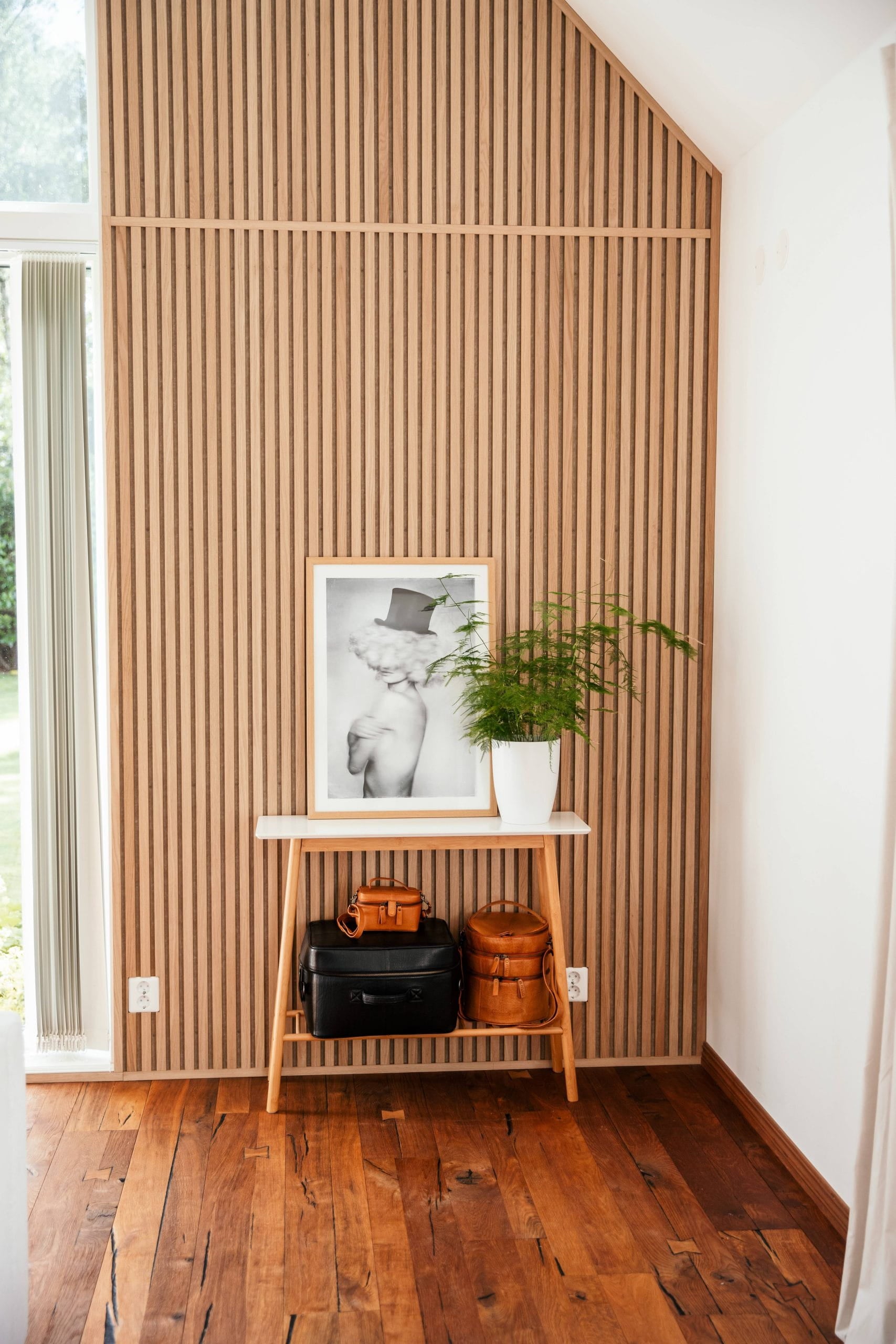 Ein kleiner Holztisch mit Taschen, einem gerahmten Foto und einer Topfpflanze steht vor einer nordischen Akustik-Lamellenwand in einem hellen Raum mit Holzboden.