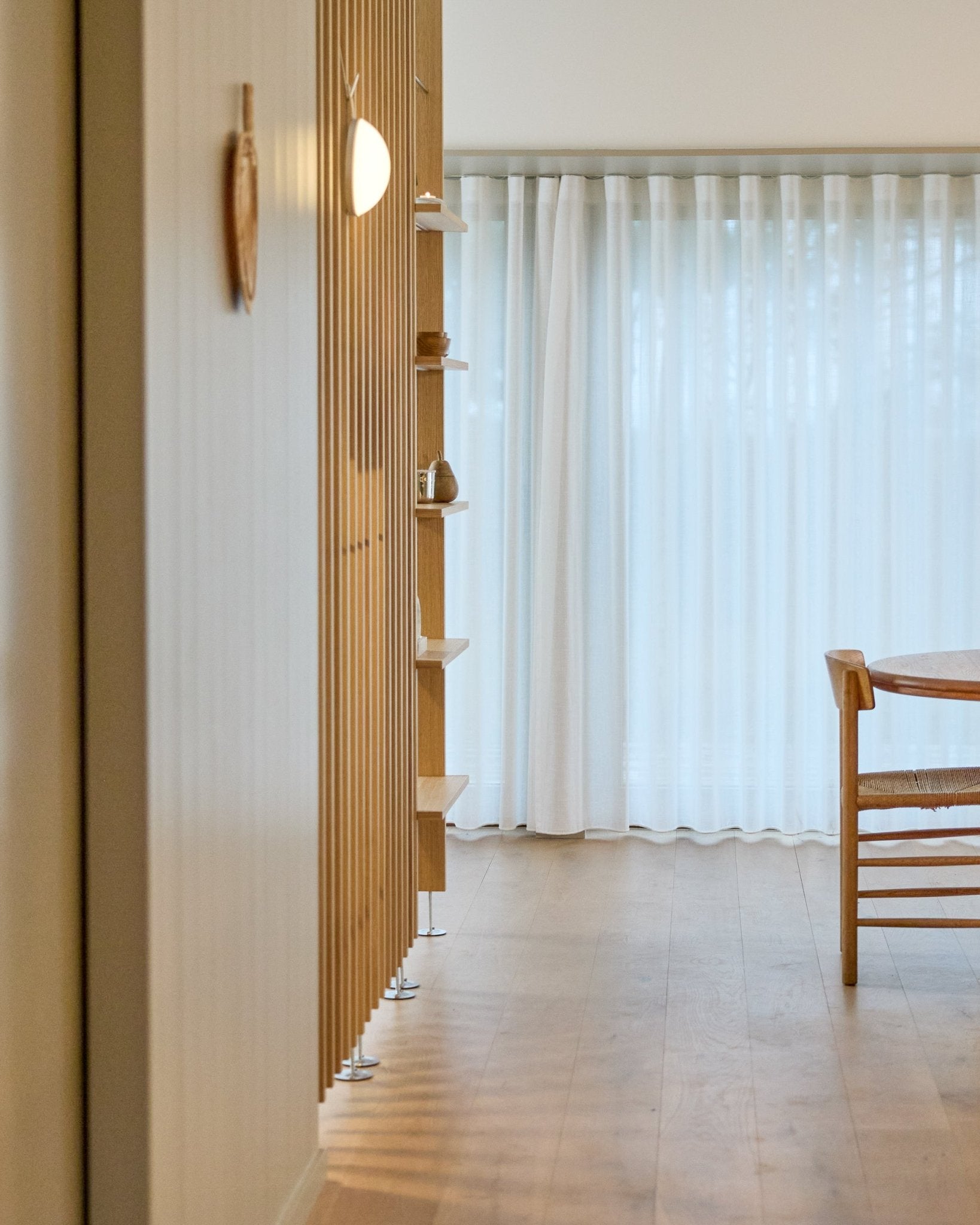 Minimalistischer Raum mit einem raumhohen WoodUpp Embrace Raumteiler, Holzboden, Regalböden, Stuhl und transparenten Vorhängen vor large Fenstern.
