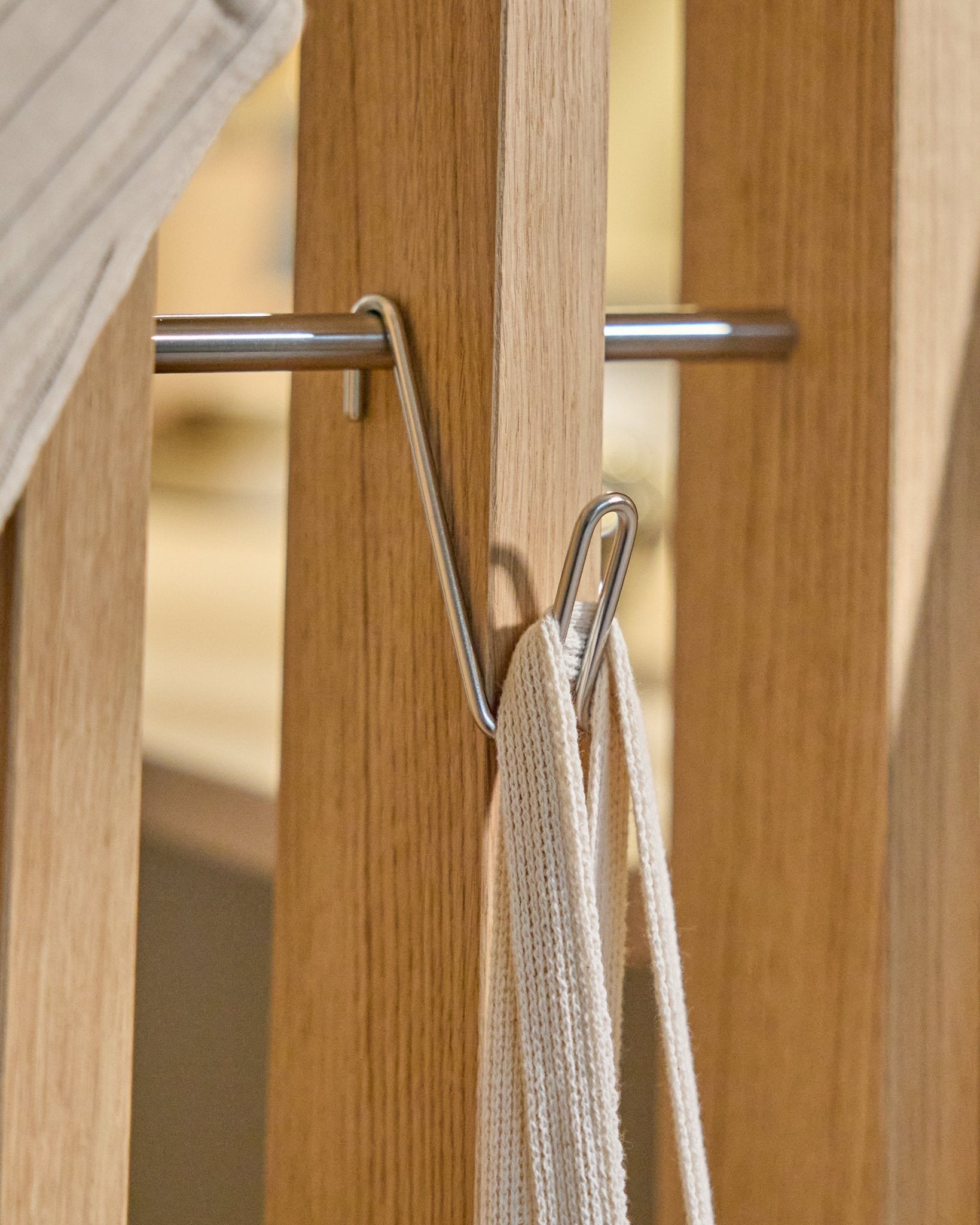 Der Alva Wire Hook / BM1 (Edelstahl Steel, kurz) von WoodUpp ist an einer Holzschiene Akustikpaneel montiert und hält eine weiße Schlaufe mit Metallstab; Hintergrund beige.