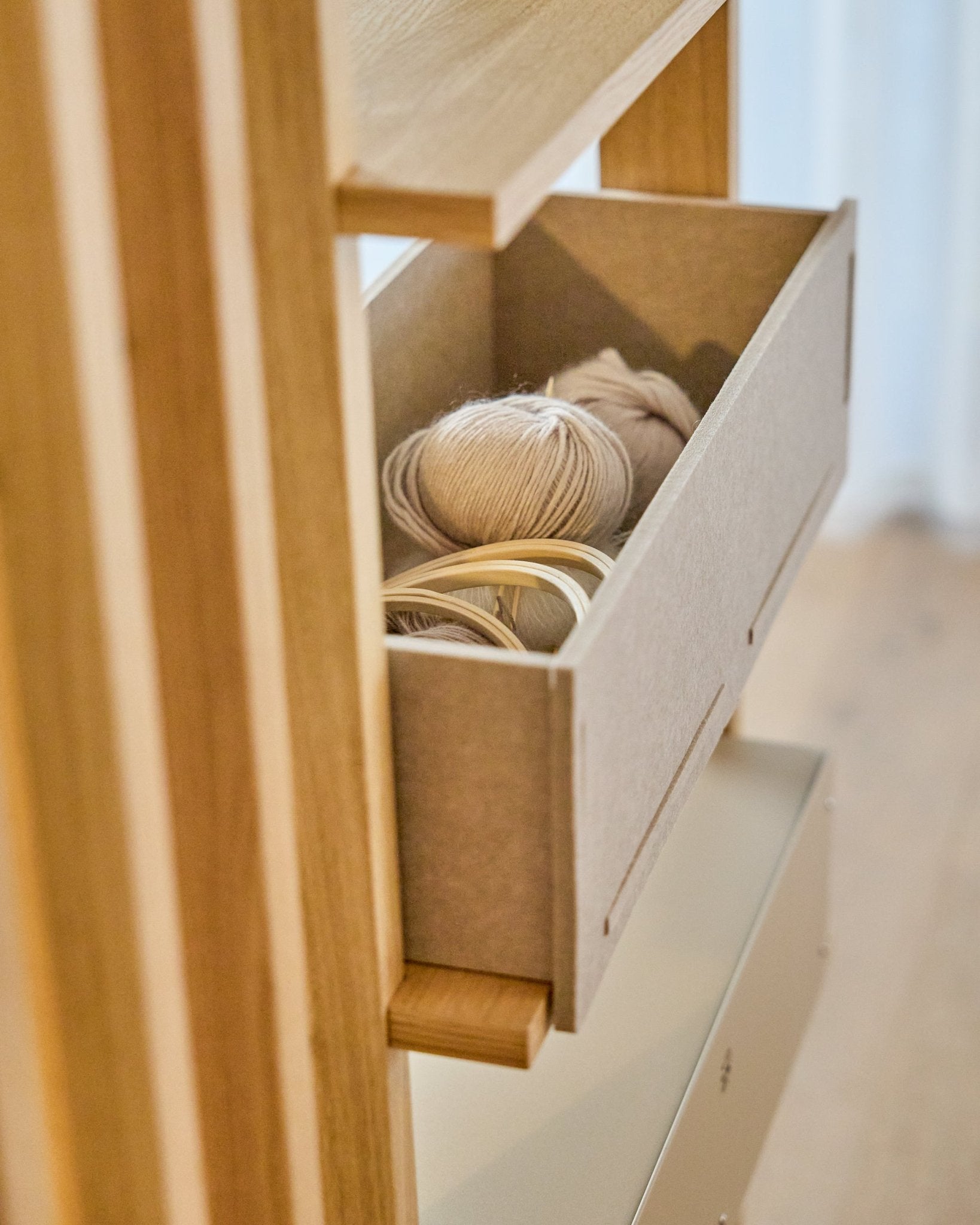 Die WoodUpp Vigg Felt Box / BM2 in beige bewahrt leichte Wollknäuel und hölzerne Stickrahmen auf einem Holzregal auf und ergänzt nordische Akustikpaneele.