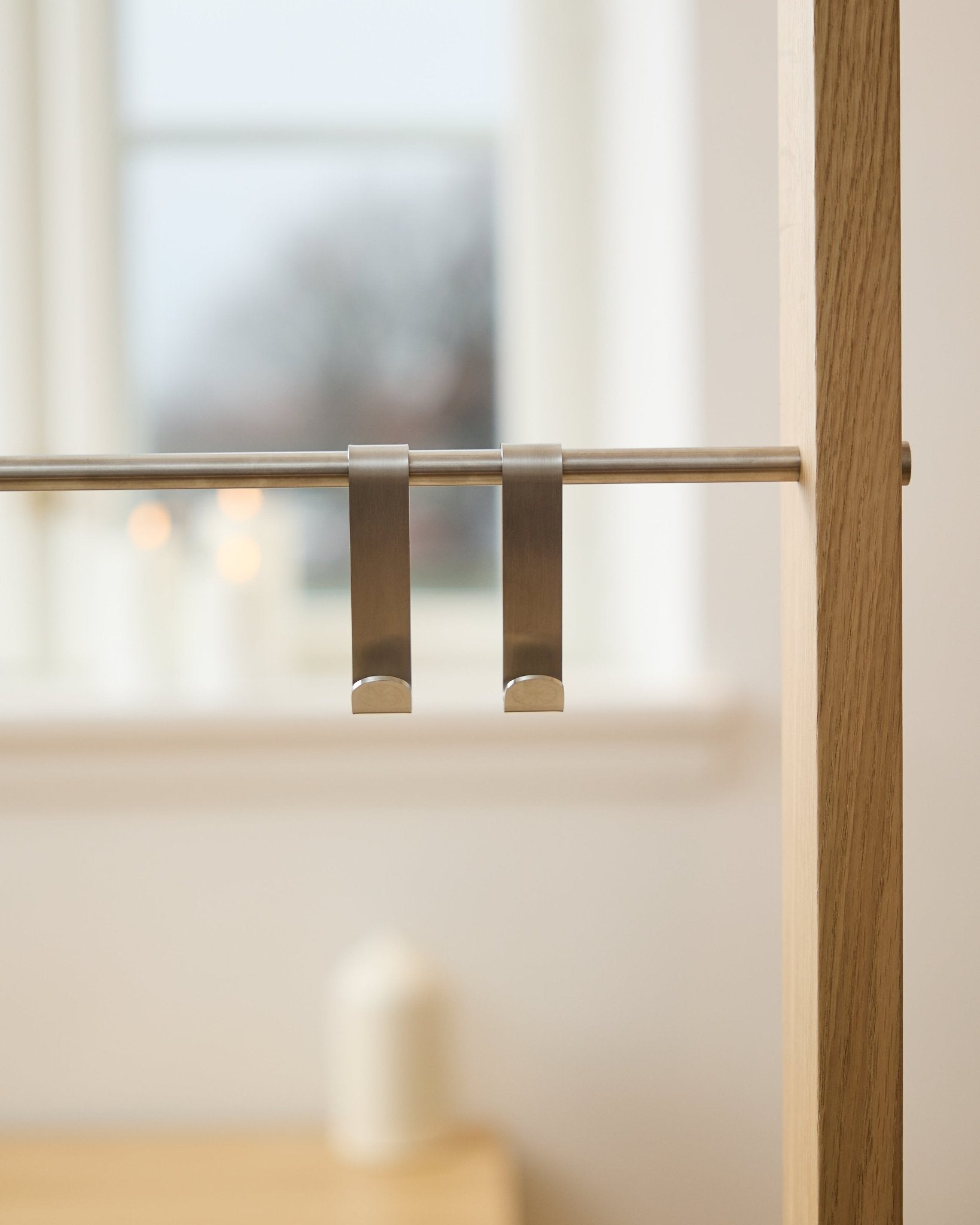 WoodUpp Alva S-Haken BM1  BM2 (2 Stk.) aus steel Edelstahl, an einer Stange eines Akustik-Holzpaneels hängend; im Hintergrund ein unscharfes Fenster und brennende Kerzen.