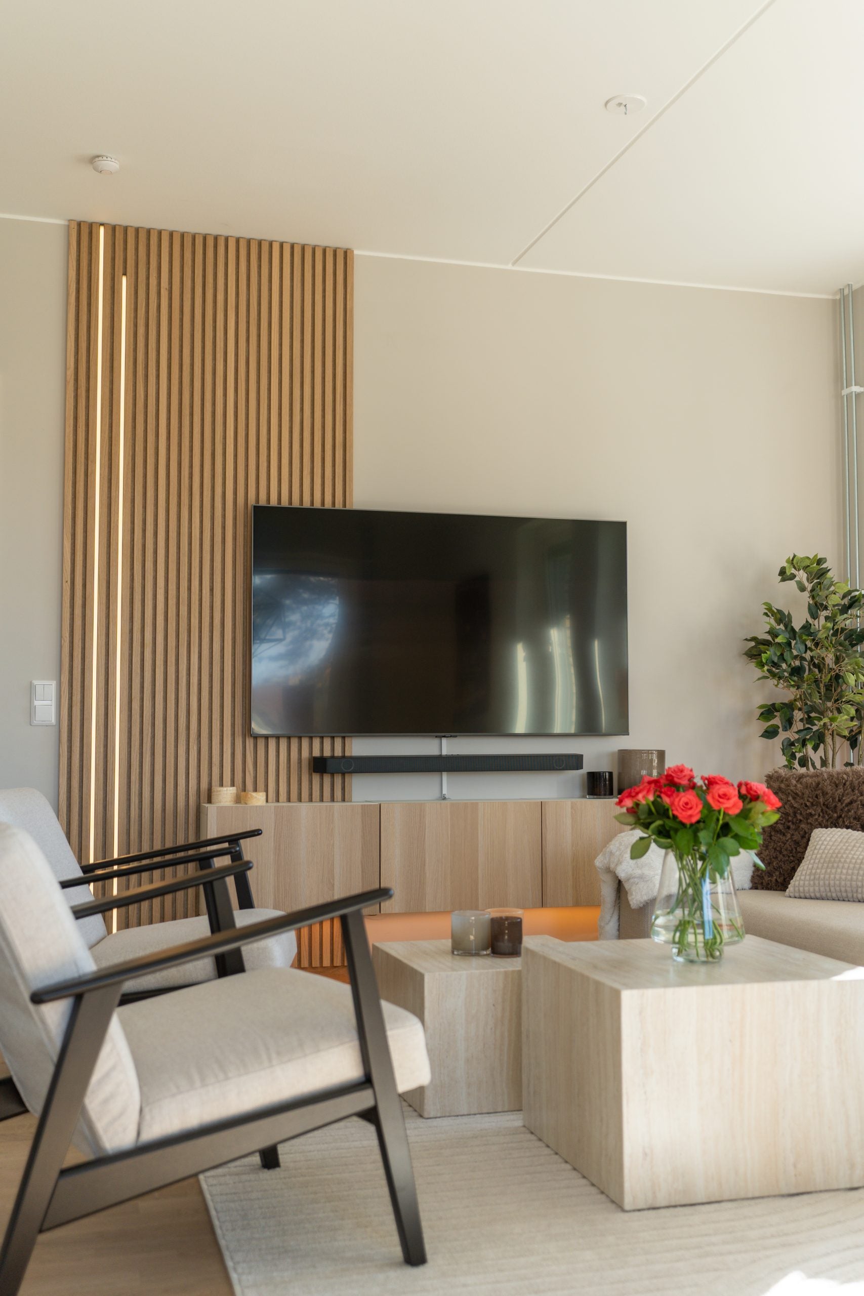 Modern living room with neutral decor, Akupanel acoustic wood paneling on the wall, flat-screen TV, armchair, coffee table, plant, candles, and red roses in a vase.