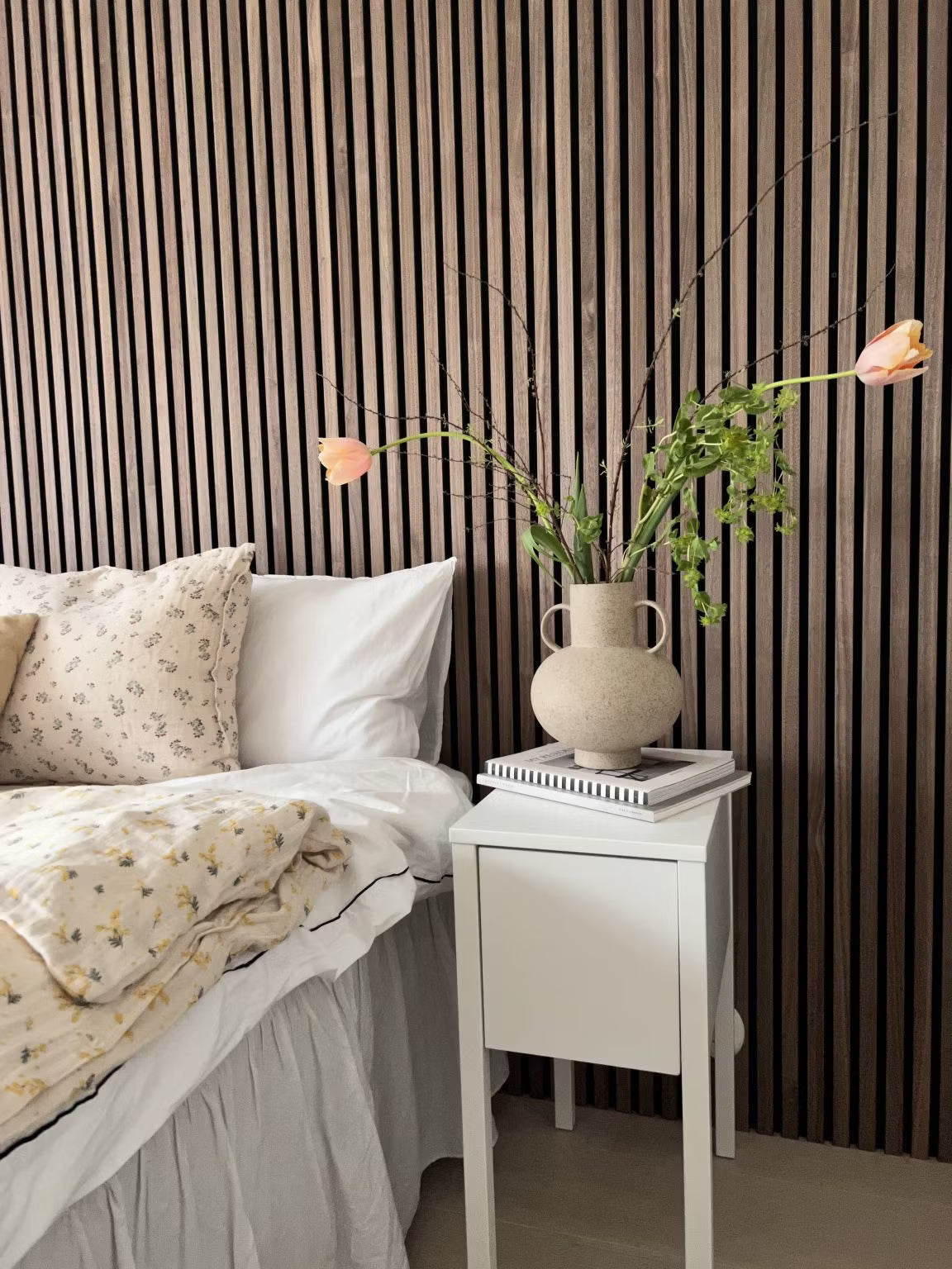 Ein Schlafzimmer mit einem weißen Bett mit geblümter Bettwäsche und einer Blumenvase, mit einer Akzentwand aus nordischen Akupanel-Holzlatten hinter dem Bett.