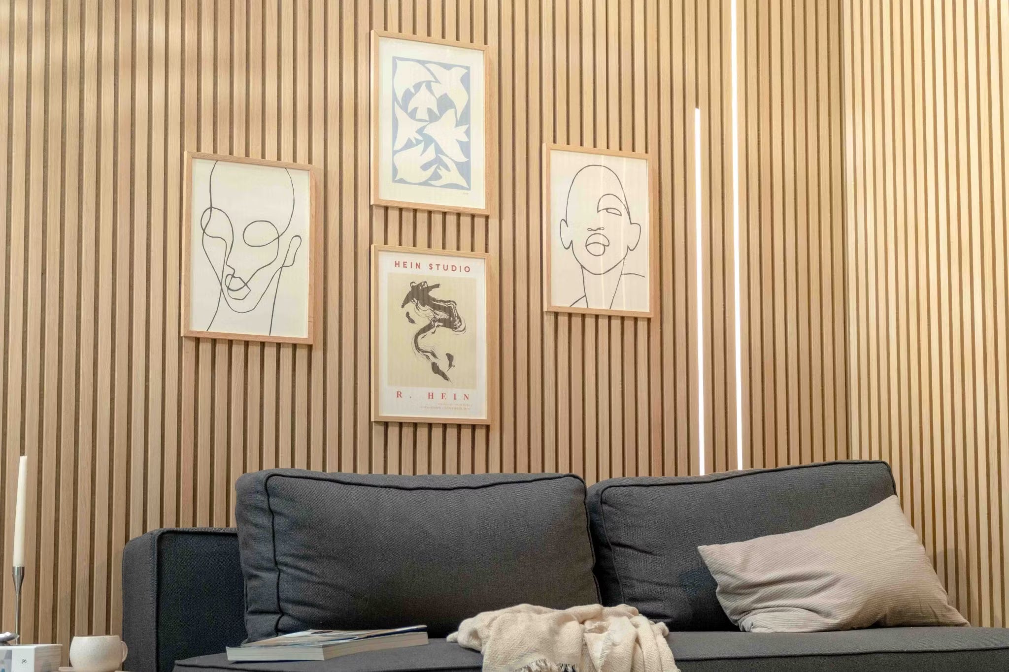 Modernes Wohnzimmer mit dunklem Sofa und beigen Kissen, akzentuiert durch eine Akupanel-Akustikholzlattenwand und minimalistische skandinavische Kunst darüber.