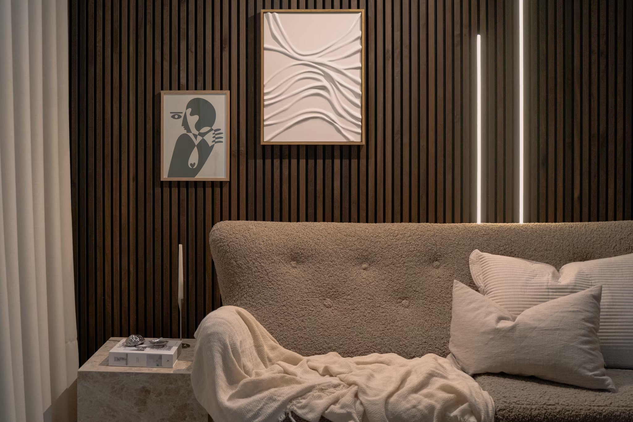 Modernes Wohnzimmer mit strukturierter beiger Couch, weißen Kissen, abstrakter Akustikpaneel-Kunst und vertikaler Akupanel-Holzlattenwand mit sanfter Raumbeleuchtung.