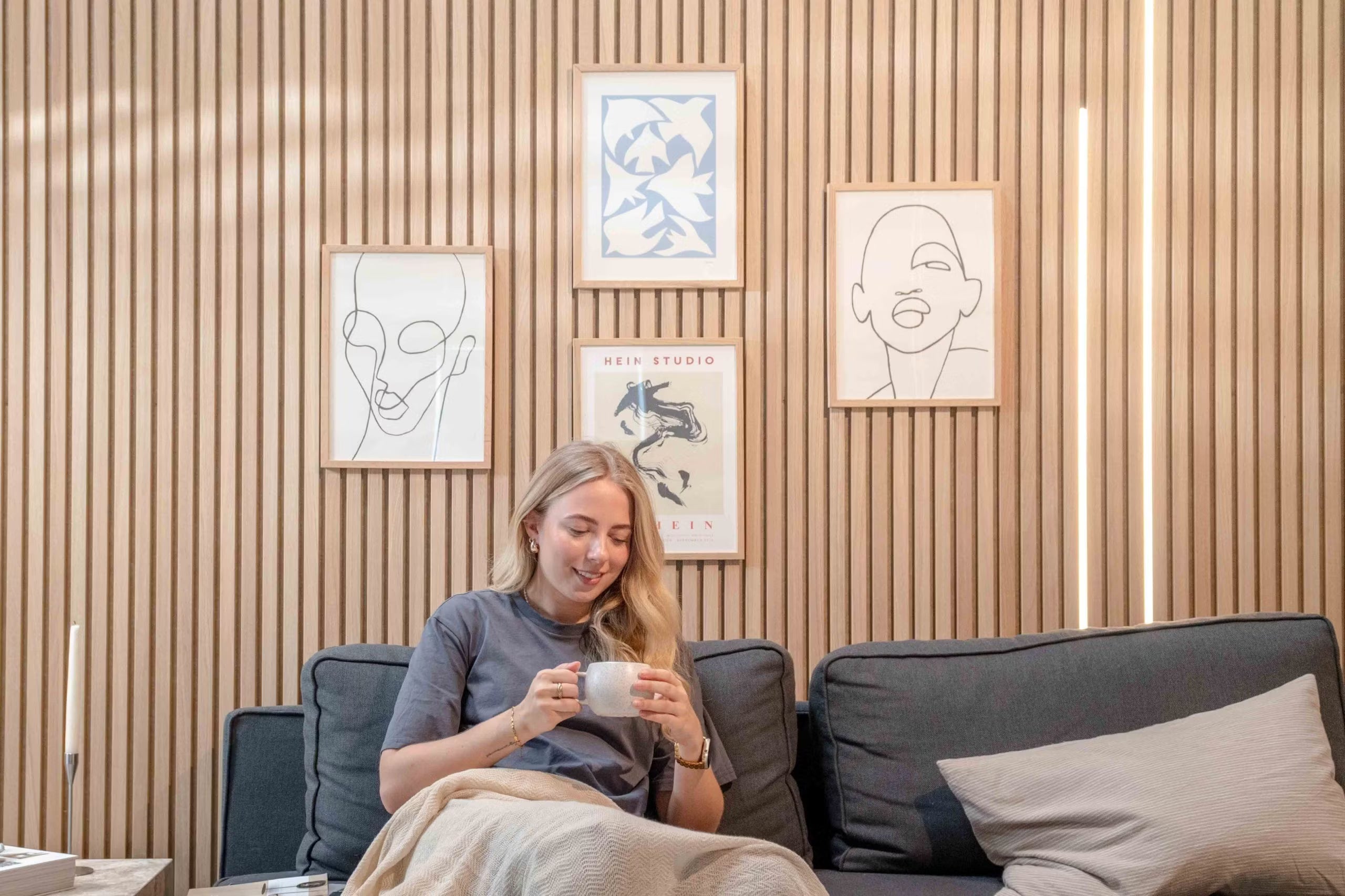 Eine Frau, die sich mit einer Tasse auf einem Sofa entspannt, lächelt unter minimalistischer Linienkunst und abstraktem Dekor, das auf strukturierten nordischen Akustik-Lamellenwänden montiert ist.