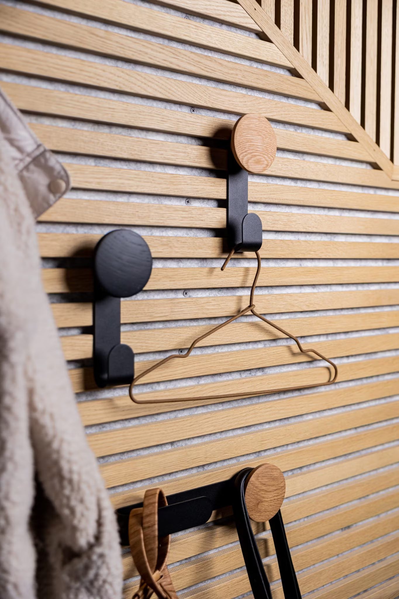Drei moderne Haken, die an einer Akupanel-Akustikholz-Lamellenwand im nordischen Stil angebracht sind, halten einen Metallbügel und Accessoires, wobei links ein wuscheliger Mantel teilweise sichtbar ist.